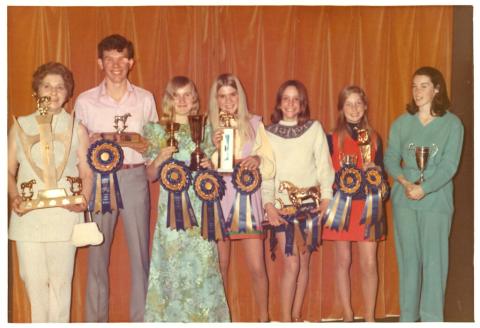 Seven young people smiling and holding trophies and ribbons.