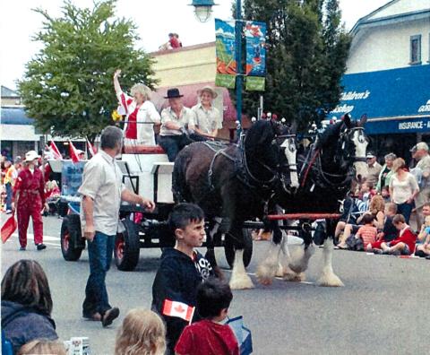 Three folks sitting on a horse-drawn carriage, moving along the centre of the road, waving at spectators.