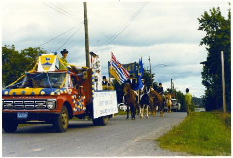 A truck with a flat deck drives ahead of horses and riders. The truck is decorated in yellow and blue.