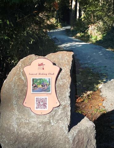 A rock in the foreground with a small sign that says Sunset Riding Club with a picture of horses and riders. Behind the rock is a gravel trail that disappears into the forest beyond.