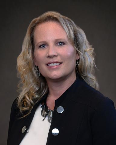 A professional headshot featuring a person with wavy blonde hair, wearing a dark blazer and necklace.