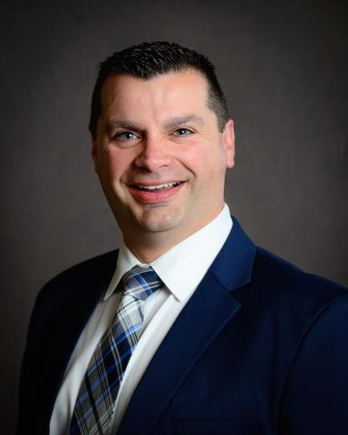 Professional portrait of a person smiling, wearing a suit and tie against a dark background.
