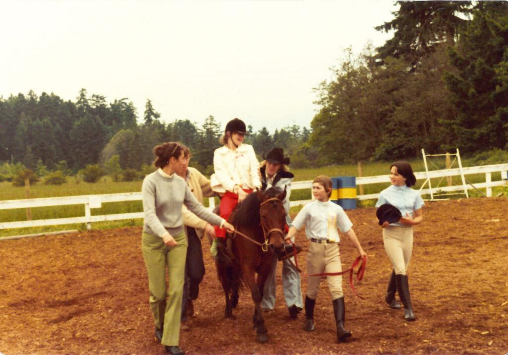 A young woman riding a pony. Walking beside her are several other young riders in riding gear, walking in a horse-riding corral.