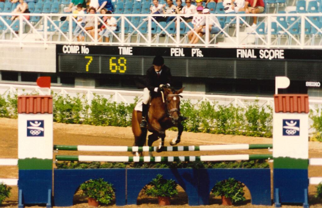 Background shows a horse race stadium. Midground shows a horse and rider jumping over a vertical jump with two poles. Looks intense.
