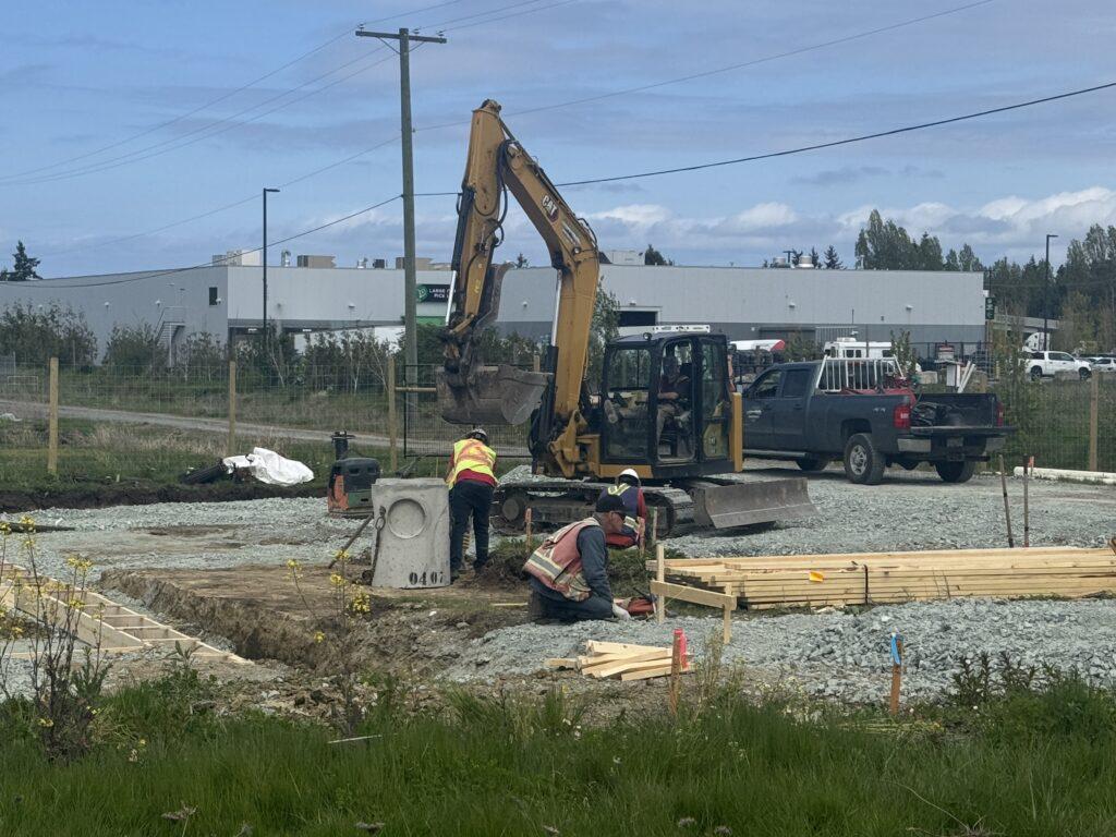 Construction workers in safety vests operating near an excavator at a gravel-filled building site.